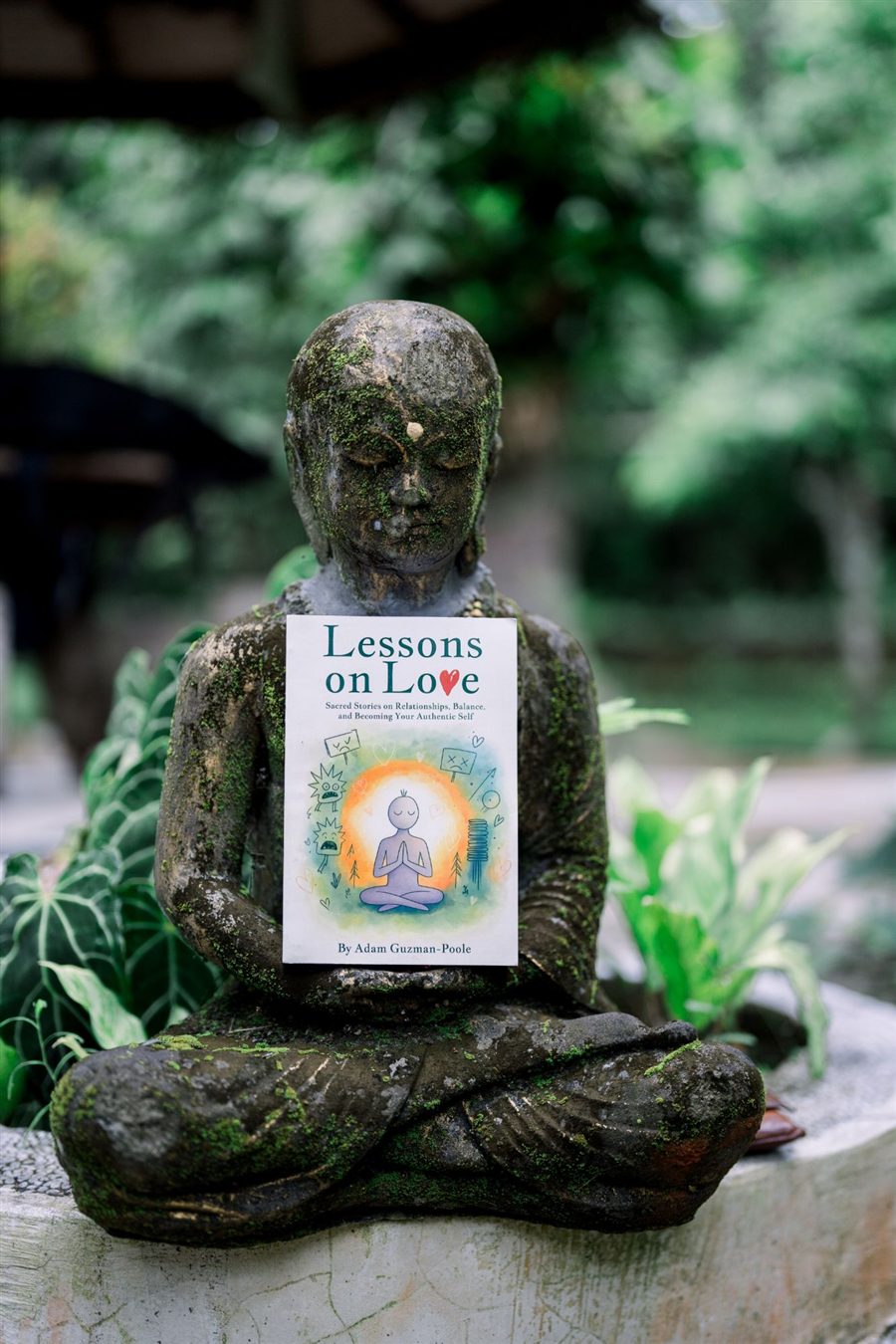 The Lessons on Love paperback held in the hands of a moss-covered Buddha statue in a lush garden