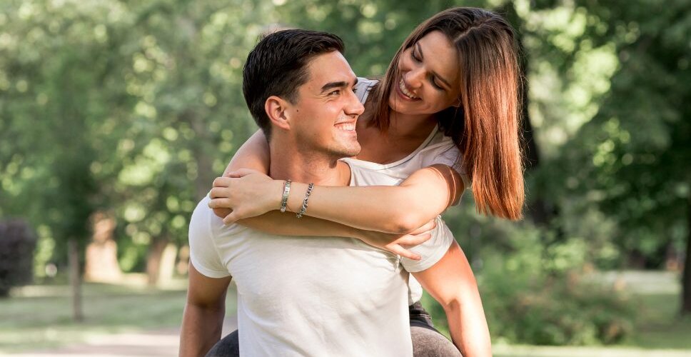 A couple laughing together in a sunny park, her arms around his shoulders